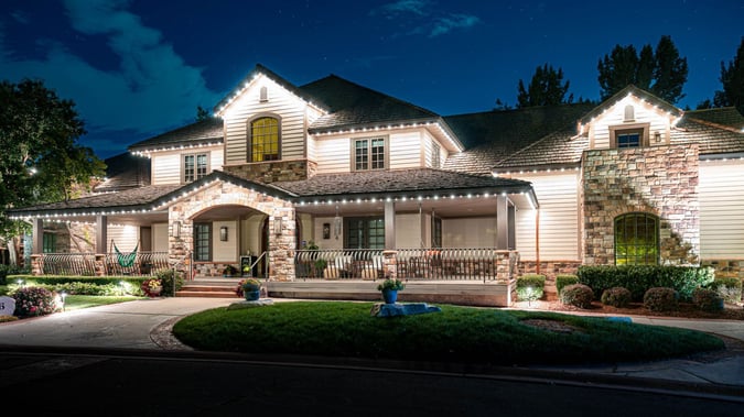 A bright home with brick rock walls and a Trimlight accent lit pattern. it has many plant vases in front and a hammock.