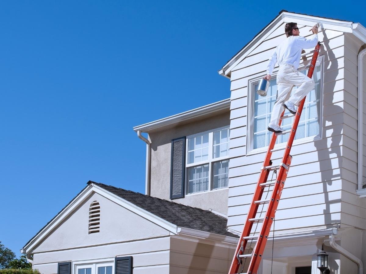 A picture of a man on a white house, painting the trim of the home while on a orange ladder.