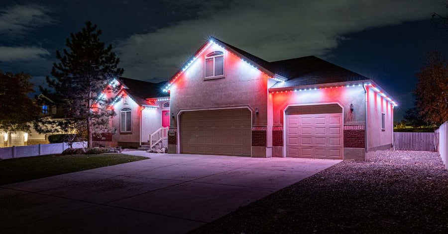 Red and white holiday lighting underneath roofline of stucco home with pine tree out front.