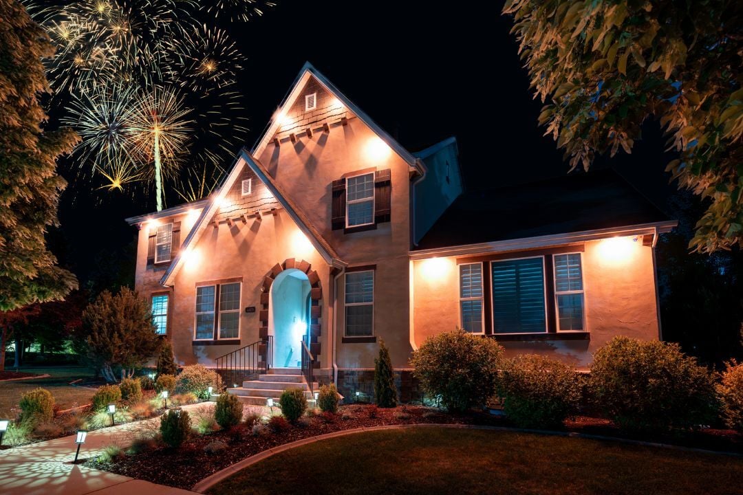 A picture of a home with downlight accent lighting that has fireworks behind the house and trees and landscape lighting out front.