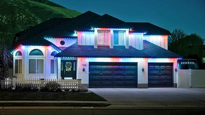 A white wide boxy home with a fence in front, and has a red, white and blue trimlight pattern.