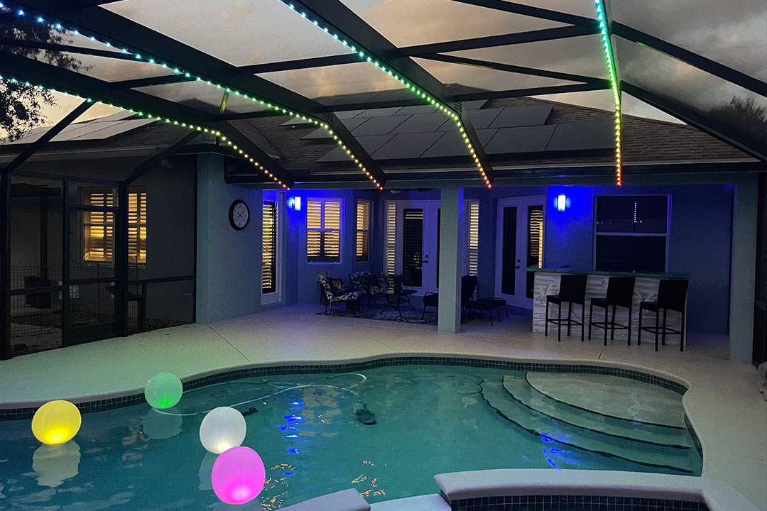 A pool cage with green and blue lights on the ridge and colorful lit up balls in the pool.