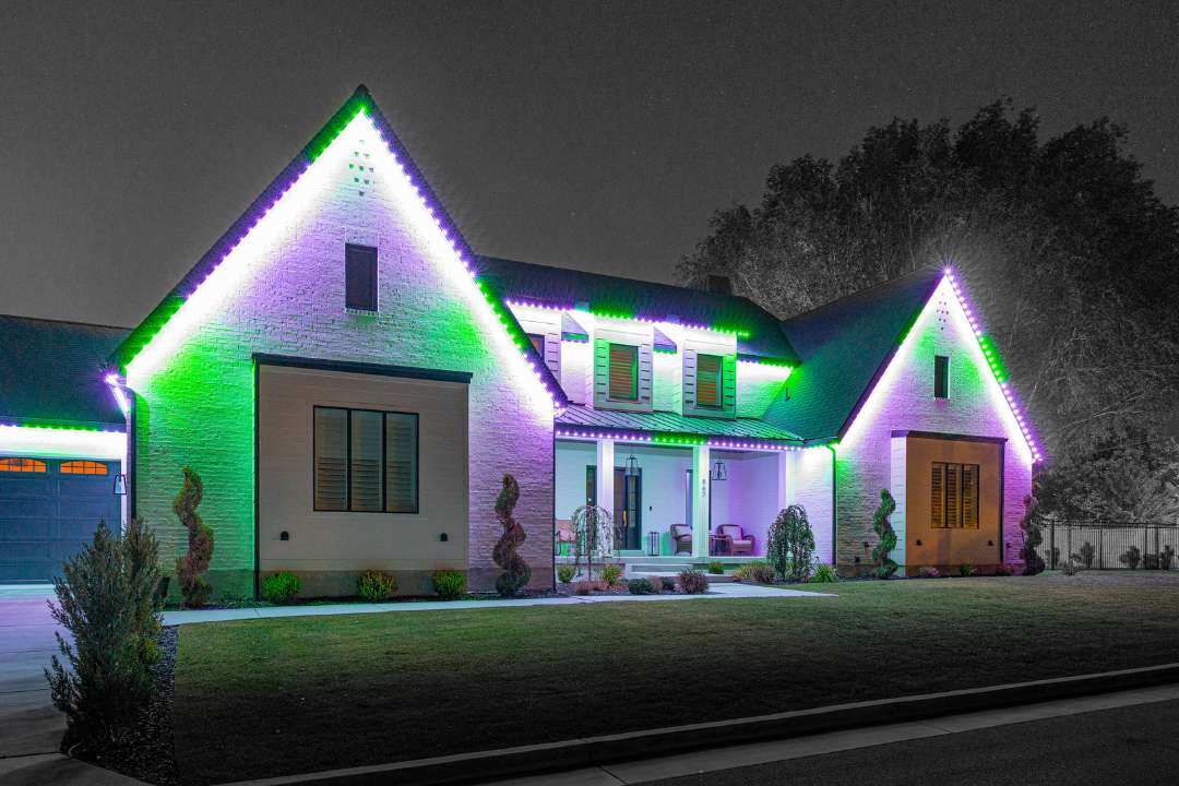 a home that has a green, purple and white lighting pattern with trees and a lawn.